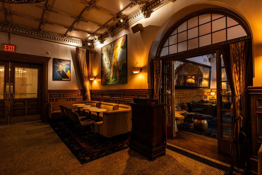 Wood-paneled rooms of Hotel Chelsea's Lobby Bar, art on the walls and an arched doorway glowing beyond