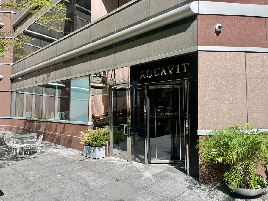 The polished glass entrance of Aquavit beneath gold lettering, flanked by potted palms and a small terrace on East 55th Street.