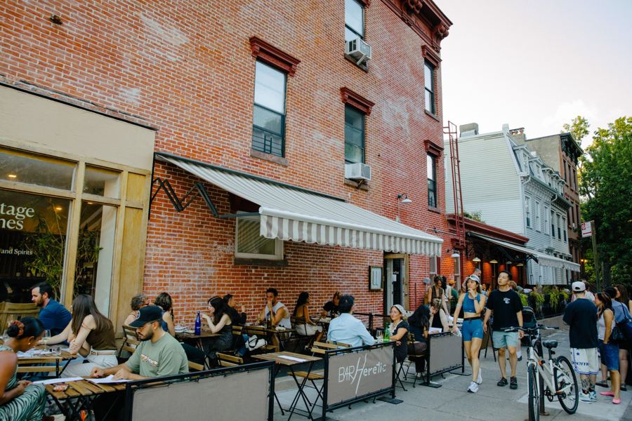 Bar Heretic's sidewalk tables full on a summer evening on Clermont Avenue in Fort Greene, under a striped awning on a brick corner building
