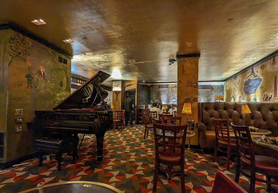 The interior of Bemelmans Bar at The Carlyle, with a black grand piano, small lamp-lit tables, a patterned carpet, a gold-leaf ceiling and Ludwig Bemelmans' painted murals on the walls.