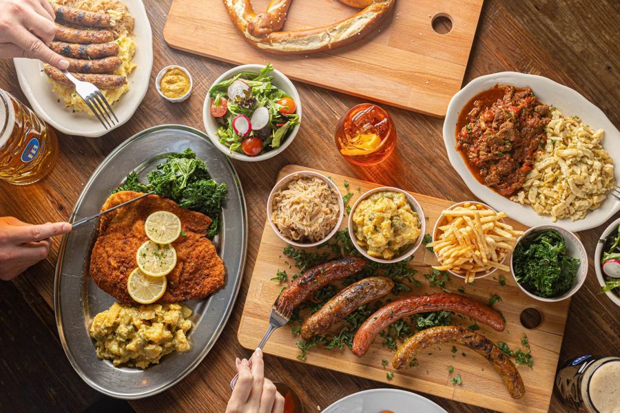 A German feast at Black Forest Brooklyn — schnitzel with lemon, grilled sausages, a giant pretzel, spätzle and a mug of beer on wooden tables