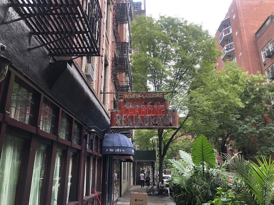 Carbone's red neon 'Wines Liquors' restaurant sign hangs above the brick facade and blue No. 181 awning on leafy Thompson Street.