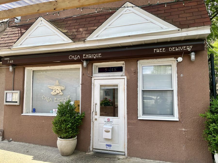 The small brown storefront of Casa Enrique in Long Island City, its name painted on the awning above a white door and a potted shrub.