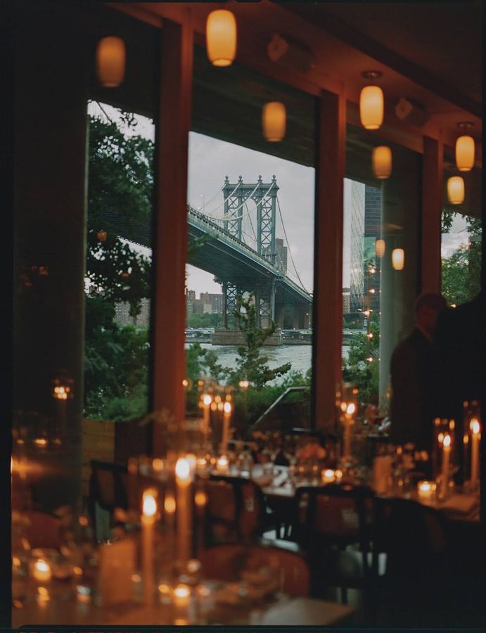 The Manhattan Bridge framed by Celestine's tall windows at dusk, with candlelit tables inside