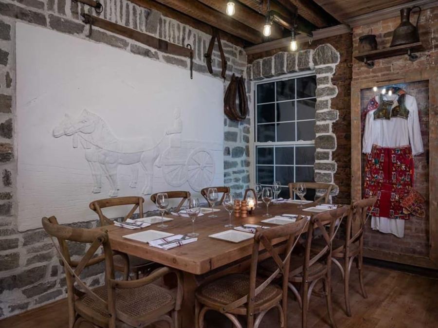 A long wooden table set for dinner under timber beams at Çka Ka Qëllu, with a horse-and-cart relief on the stone wall and a traditional Albanian costume on display