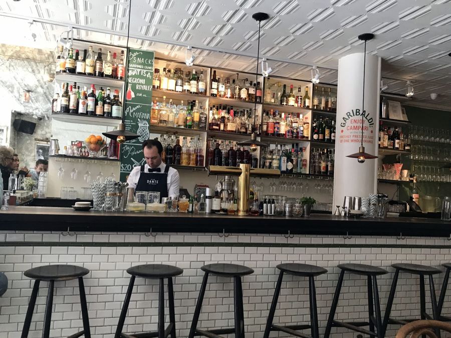 A bartender works behind Dante's white-tiled bar, with stools in front and backlit shelves of bottles under a pressed-tin ceiling.