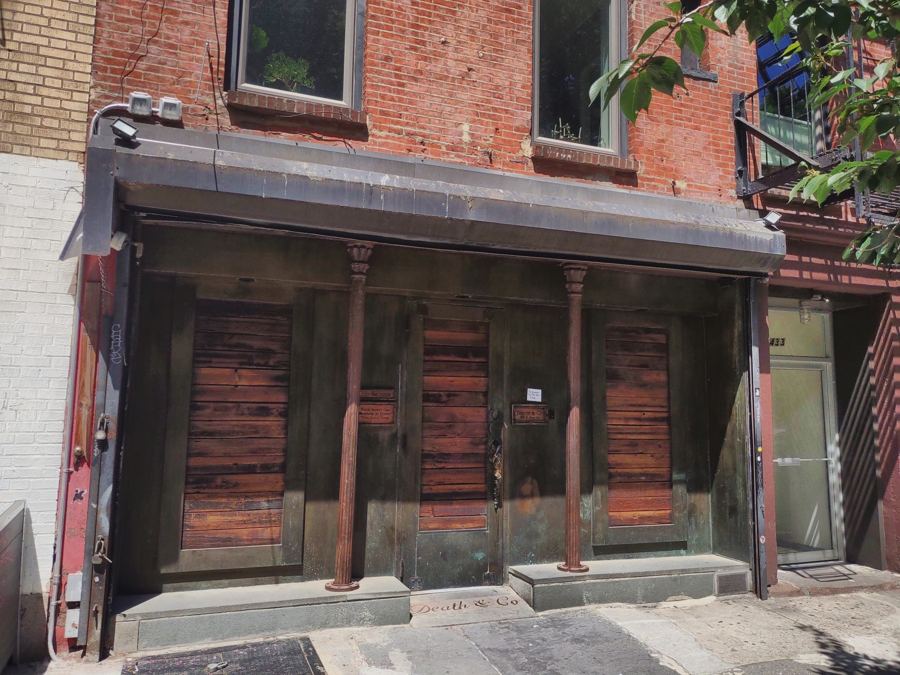 The unmarked, weathered wooden doors and iron columns of Death & Co on East 6th Street, closed up on a sunny afternoon, with the bar's name inlaid in the sidewalk.