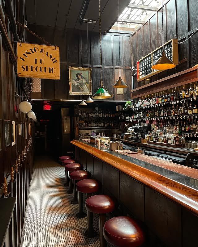 The long wooden bar at Dutch Kills under a skylight, with red leather stools, hanging pendant lamps and shelves of bottles