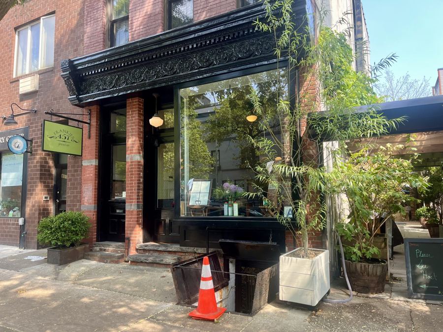 The brick storefront of Frankies 457 Spuntino on Court Street, with an ornate black cornice, a green hanging sign and leafy plants by the entrance.