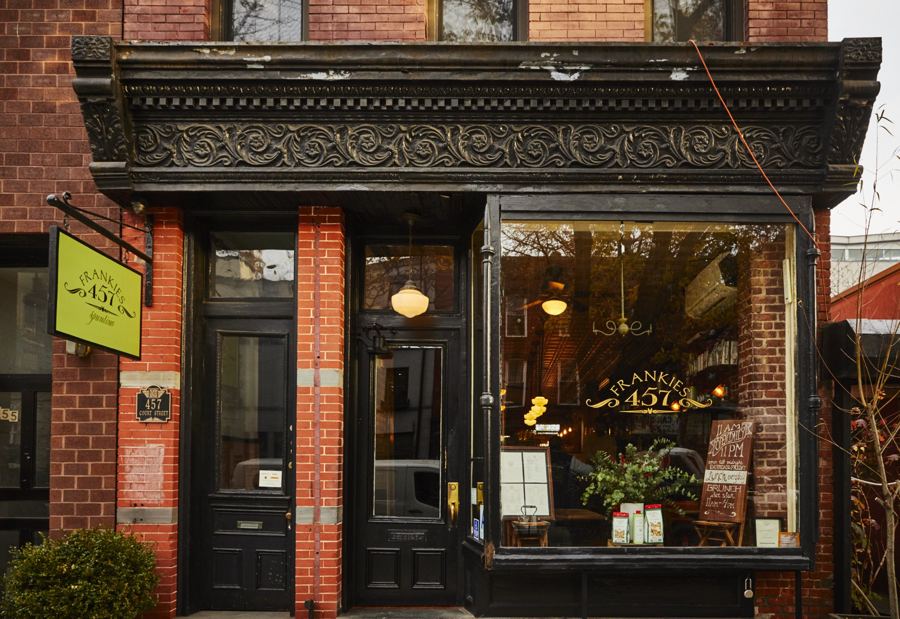 The brick storefront of Frankies 457 Spuntino on Court Street, with an ornate black cornice, a green hanging sign and leafy plants by the entrance.
