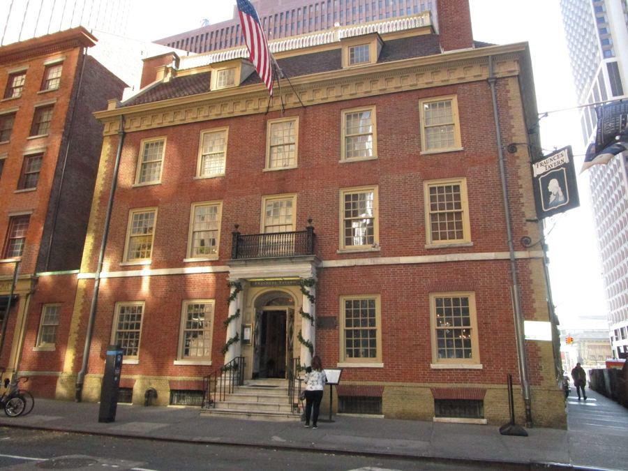 The red-brick Georgian façade of Fraunces Tavern, with its hanging portrait sign and American flag, on a sunny morning at Pearl and Broad Streets in the Financial District.