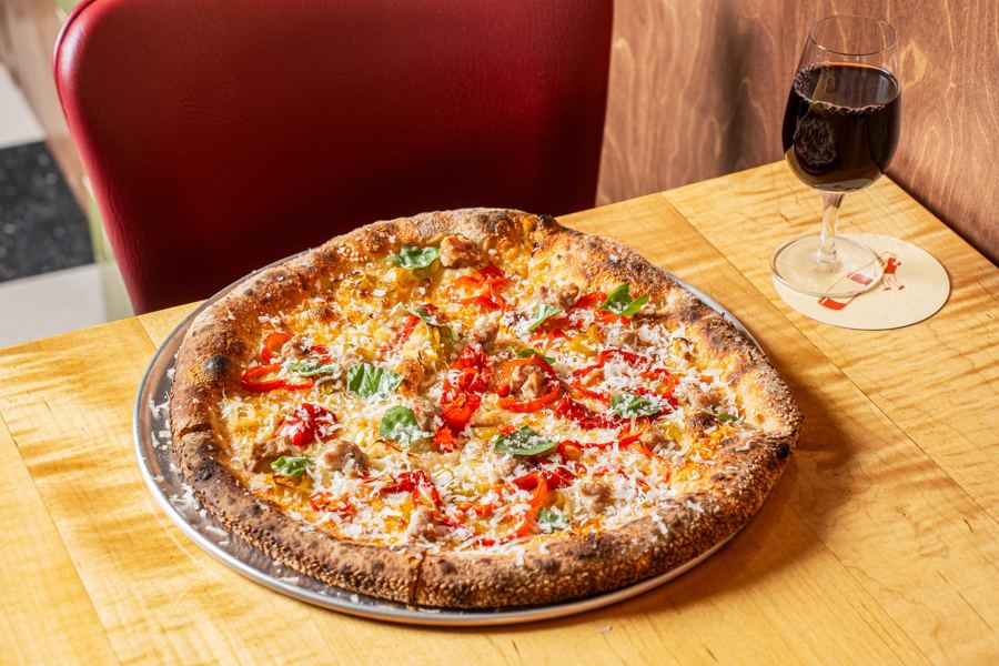 A sausage and peppers pizza on a wooden table with a glass of red wine