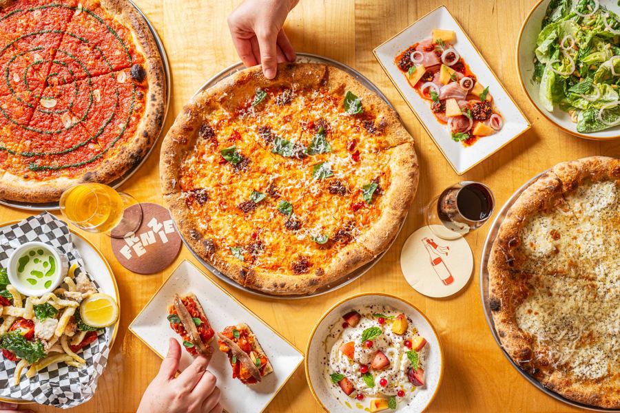 A spread of Frenzie's pizzas, salads and small plates covering a sunny wooden table