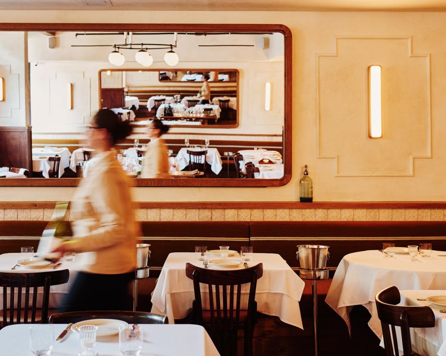 A server passing white-clothed tables in Graciela's cream-walled West Village dining room, reflected in a long mirror