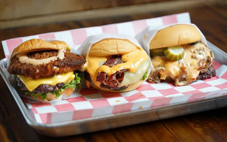 Three Halalbee's burgers in paper wraps on a red-and-white checkered tray, dripping with cheese and sauce