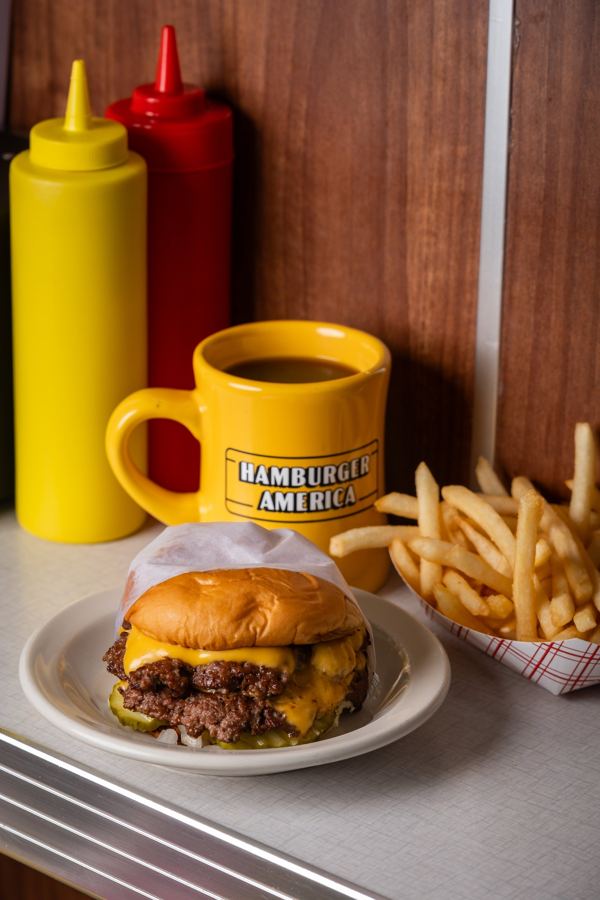 A fried onion burger with melted American cheese on a paper wrapper at a chrome diner counter, a glass of lemonade beside it