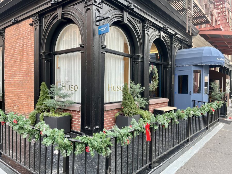 The corner of a black cast-iron storefront with 'Huso' lettered in gold on arched windows and a blue Marky's awning on Greenwich Street, Tribeca