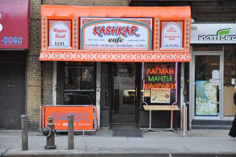 The orange patterned awning of Kashkar Cafe on Brighton Beach Avenue, with Uyghur Food signage and neon Cyrillic window signs for lagman, manty and shashlik