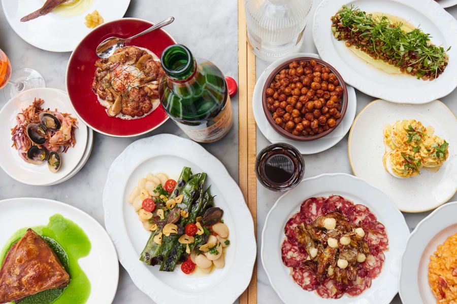 A marble table covered in La Vara small plates seen from above: cured meats, chickpeas, clams, grilled greens over white beans and a wedge of crisp roast meat, with wine
