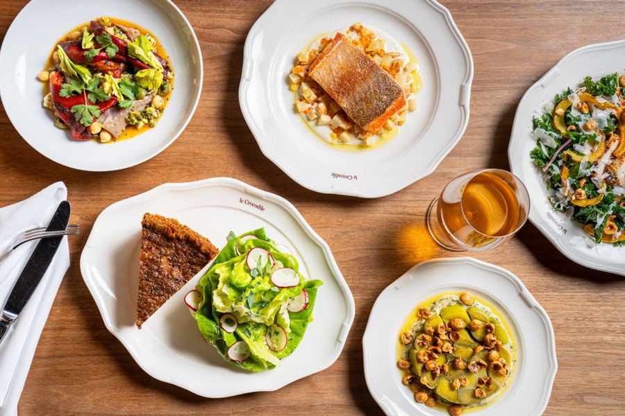 A spread of Le Crocodile's brasserie dishes on scalloped white plates: trout, salads, a slice of tart and a cocktail
