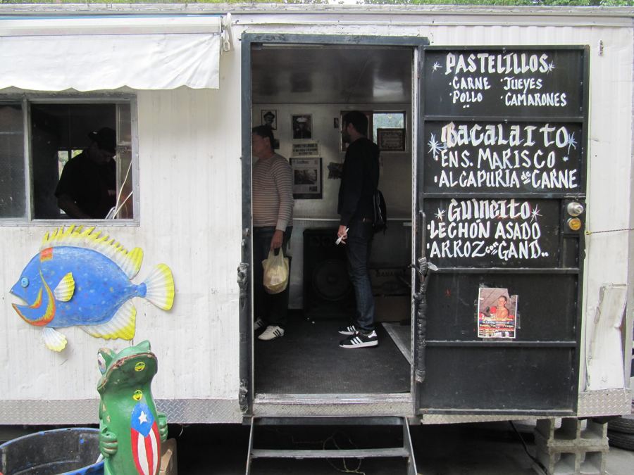 The open door of Lechonera La Piraña's white trailer, with a hand-painted chalkboard menu of pasteles, bacalaítos and lechón asado and a painted fish.