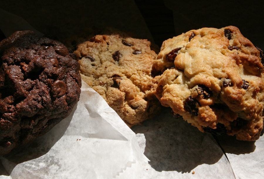 Three craggy, oversized Levain Bakery cookies — chocolate chocolate chip, oatmeal raisin and chocolate chip walnut — on wax paper.