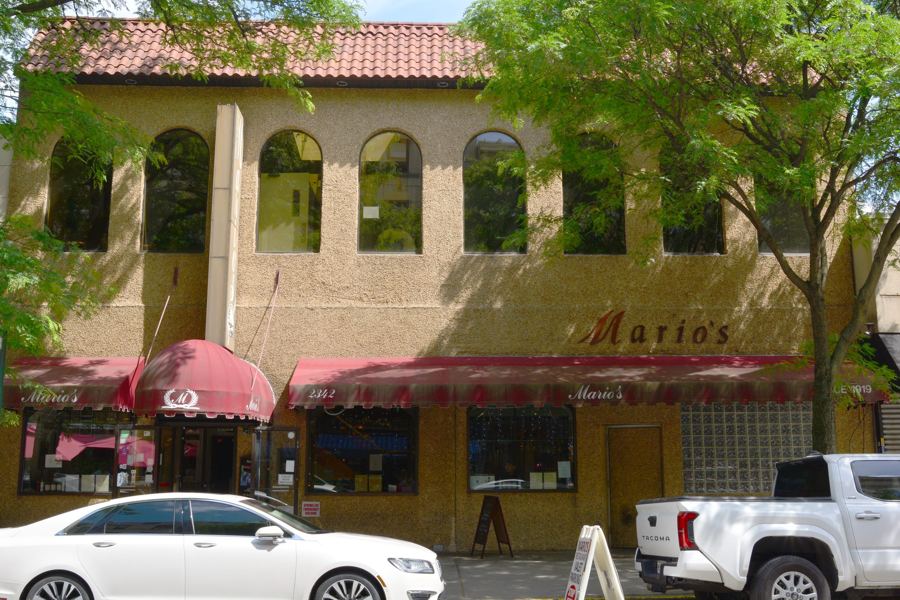 The tan stucco, tile-roofed façade of Mario's Restaurant on Arthur Avenue, with its script 'Mario's' sign and maroon awnings over the front windows on a leafy spring day.