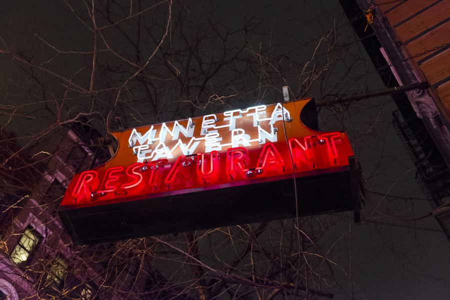 The glowing red-and-white neon Minetta Tavern Restaurant sign seen from below against the night sky and bare tree branches.