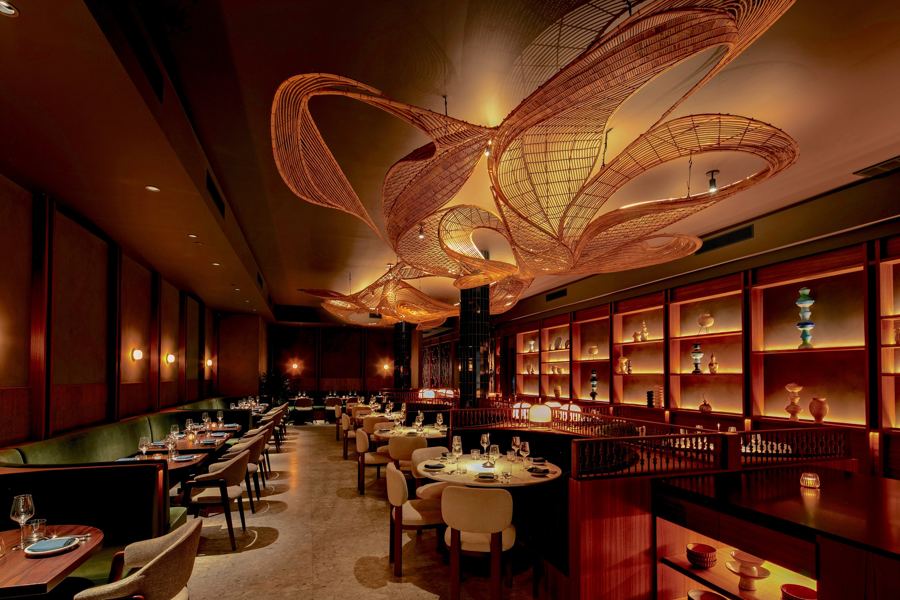 Narkara's long, warmly lit dining room under a sweeping woven ceiling sculpture, with banquettes and lit shelves along the walls