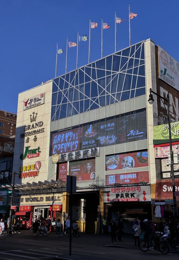 The glass-and-steel Main Street entrance of New World Mall in Flushing, layered with Chinese-language restaurant and supermarket signs under a blue sky.