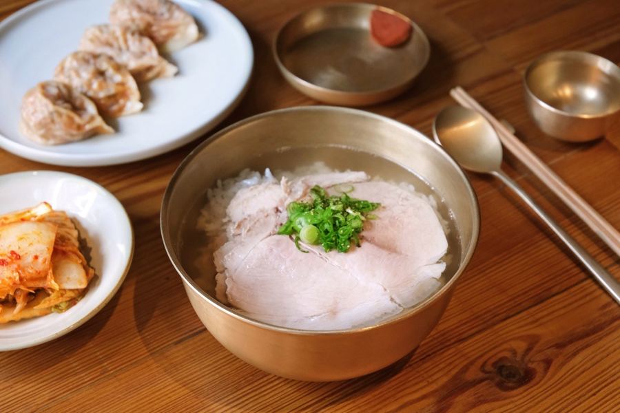 A brass bowl of clear dweji gomtang topped with sliced pork and scallions, with mandu and napa kimchi on a wooden table at Okdongsik