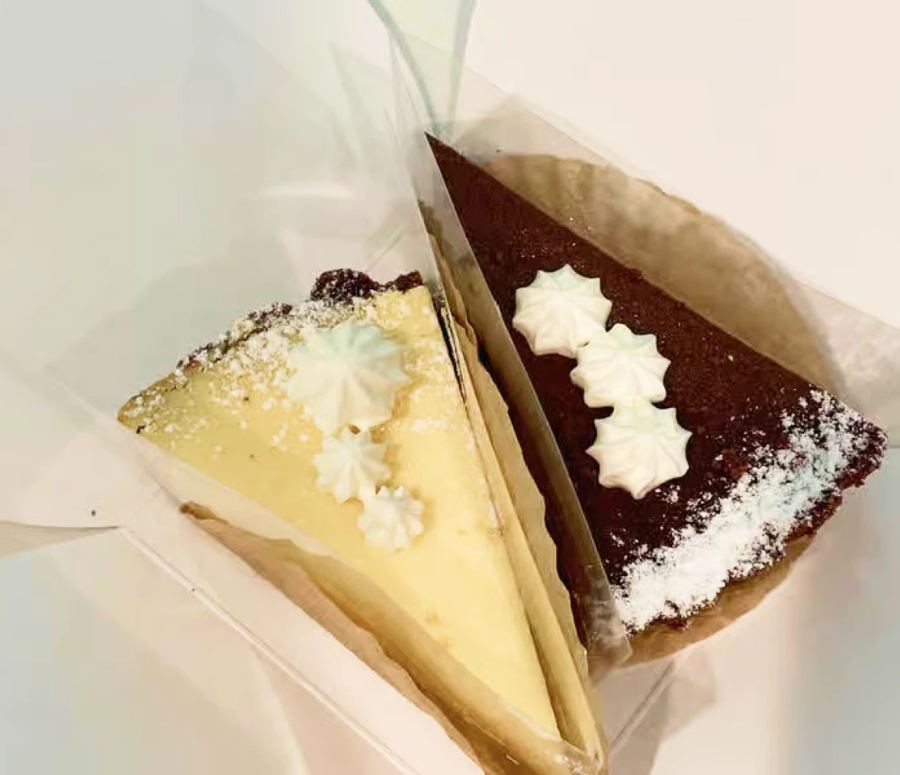 Two cake slices from Patisserie Tomoko — a pale cheesecake and a dark chocolate cake, dusted with sugar and piped meringue — in clear takeaway boxes