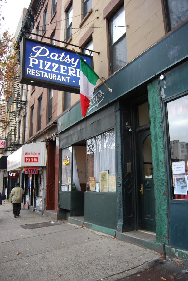 The dark green storefront of Patsy's Pizzeria in East Harlem, with its blue-and-white hanging sign and an Italian flag over the First Avenue sidewalk.