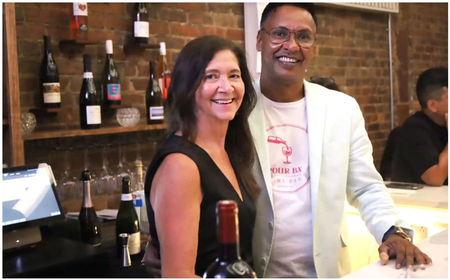Pour BX's owners smiling behind the bar, wine bottles shelved on the brick wall