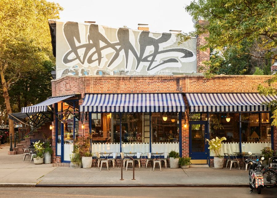 Sailor's brick corner storefront in Fort Greene under navy-and-white striped awnings, with café tables and potted hydrangeas on the sidewalk