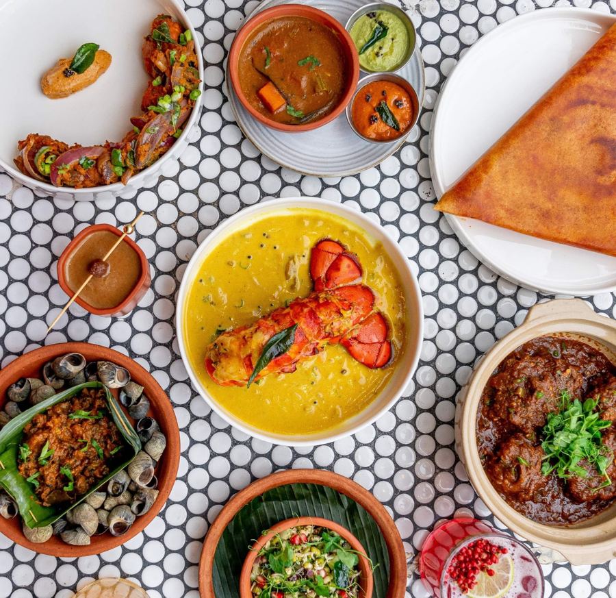 An overhead spread of Semma dishes on penny tile — lobster in a yellow curry, a dosa, snails, and clay pots of curry