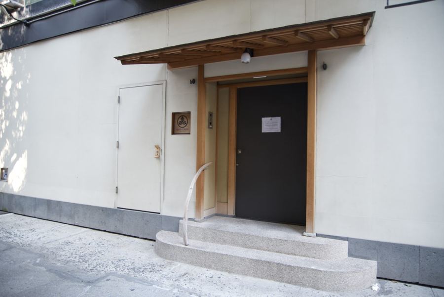 The minimalist entrance of Sushi Noz: a dark door framed in pale wood under a small timber canopy, reached by stone steps against a white wall.