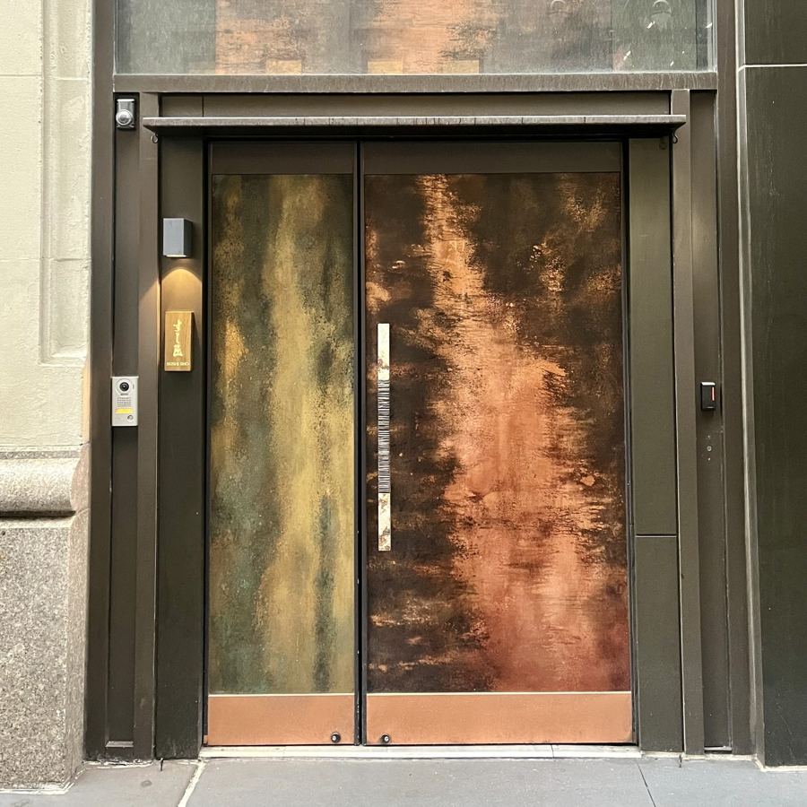 The discreet entrance to Sushi Sho in Midtown: tall patinated copper doors set into a dark steel frame beside a small brass plaque.
