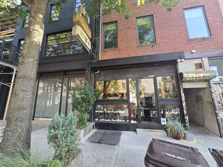 The dark storefront of The Four Horsemen wine bar on Grand Street, with wine bottles lined up in the front window and an old vintage sign hanging above