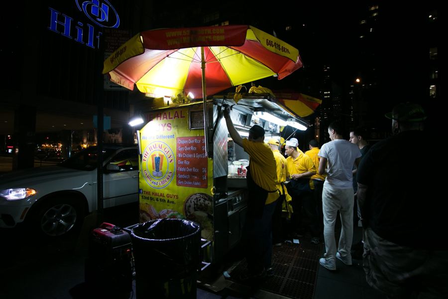 The Halal Guys' yellow menu cart glows under red-and-yellow umbrellas at night as staff in yellow shirts serve a late queue outside the Hilton in Midtown.