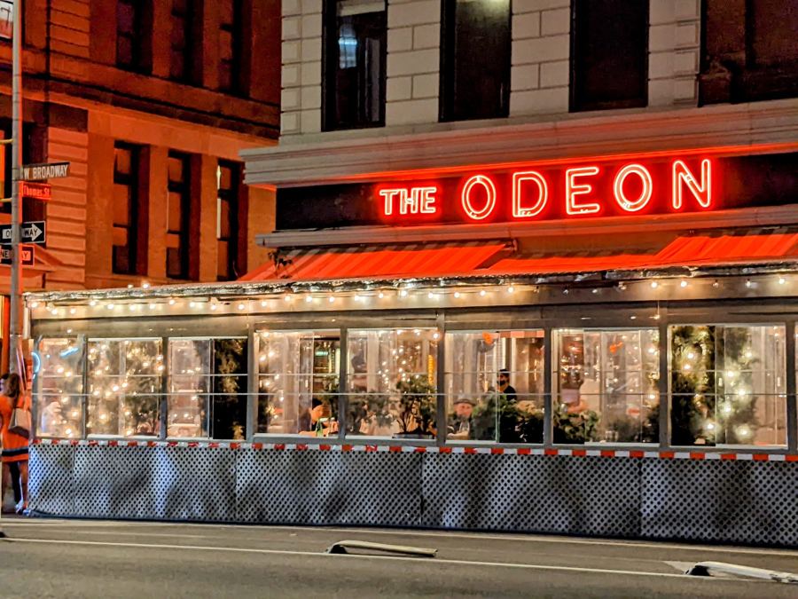 The Odeon's red neon 'The Odeon' sign and striped awning glow above its lit-up, glass-enclosed sidewalk dining room on West Broadway in Tribeca at dusk.