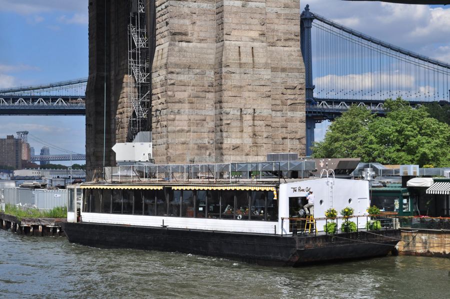 The white River Café barge, its name lettered on the wall, moored on the East River below a stone tower of the Brooklyn Bridge with the Manhattan Bridge behind.