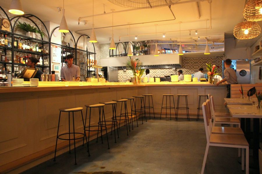 The long pale-wood bar at Two Hands in Tribeca, lined with black stools under arched shelving, pendant lamps and woven light shades.