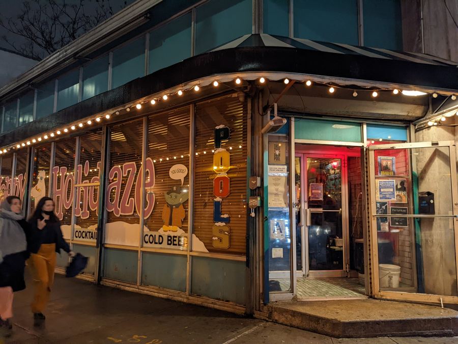 The Union Pool bar's corner storefront in Williamsburg at night, with string lights, a vertical 'POOL' sign and painted window lettering