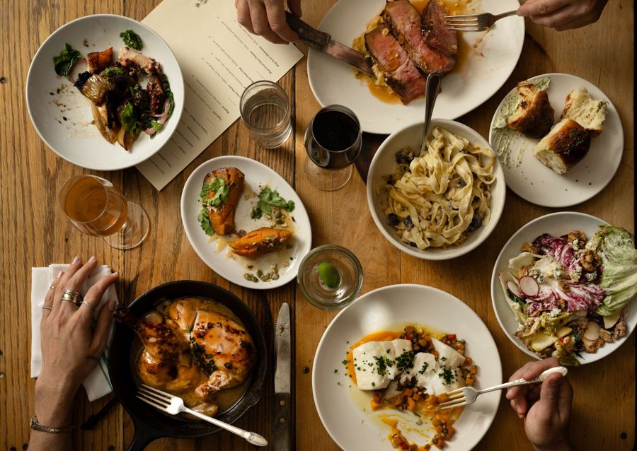 A dinner spread at Vinegar Hill House seen from above: skillet-roasted chicken, sliced steak, ribbon pasta, a chicory salad and roasted squash on a wooden table, with wine