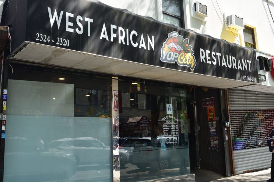 The black 'West African YopCity Restaurant' sign with its cartoon logo above wide glass windows at 2324 Arthur Avenue in the Bronx.