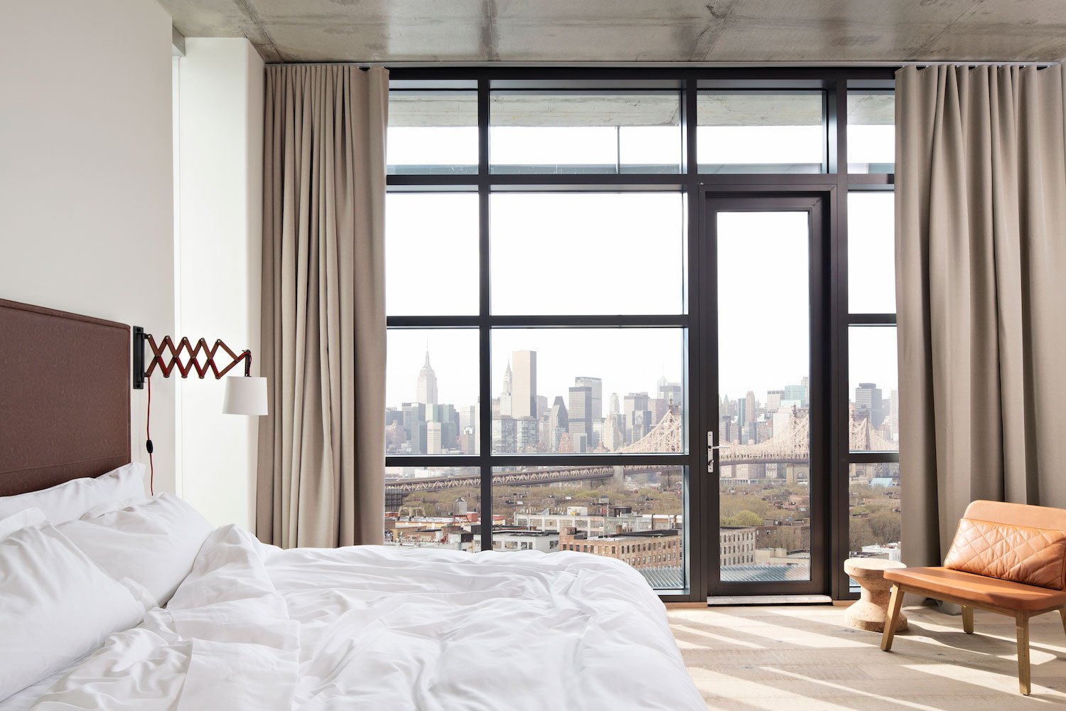 A Boro Hotel guest room with floor-to-ceiling windows framing the Manhattan skyline, a white bed and a leather daybed