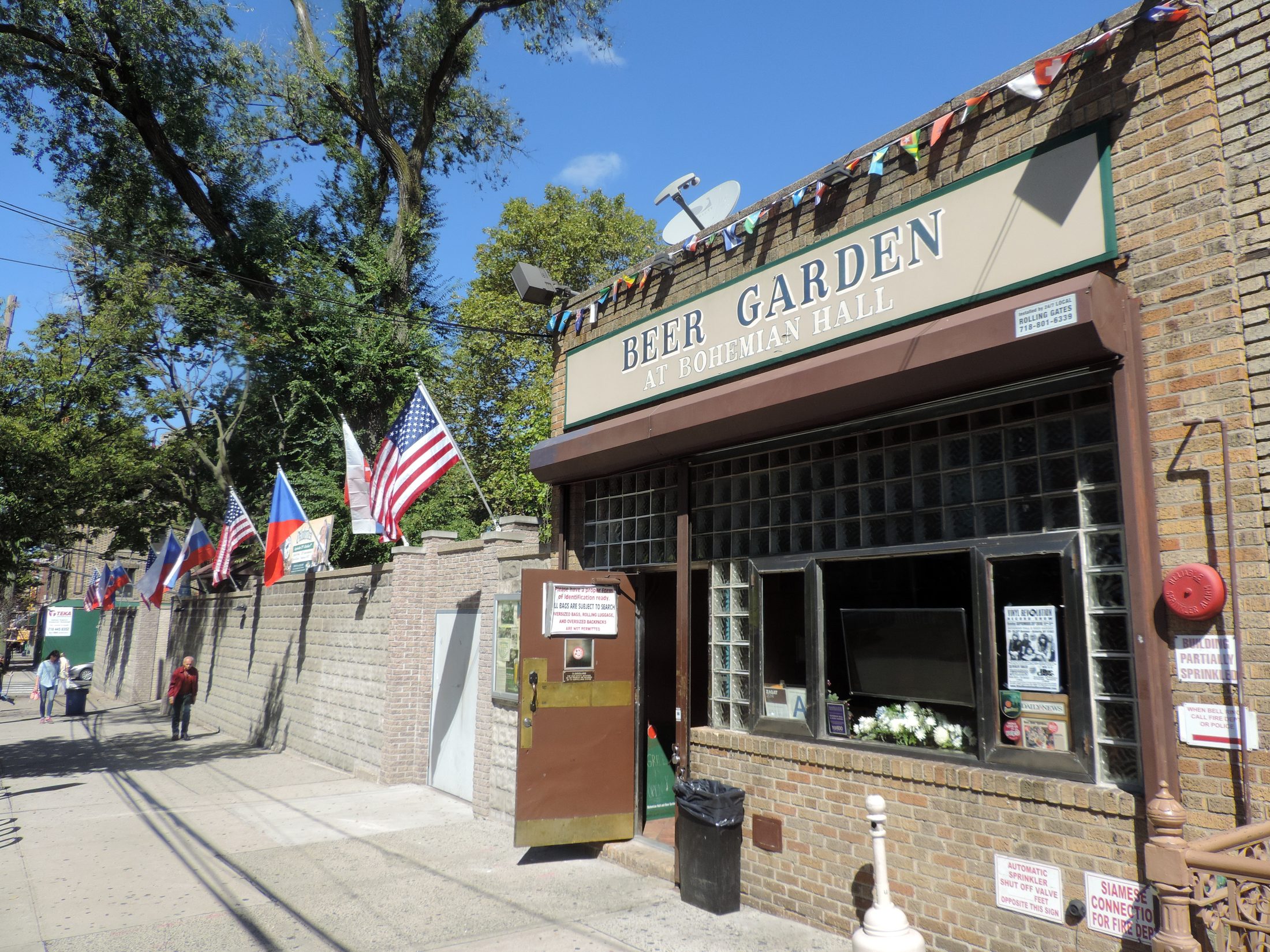 The brick entrance of the Beer Garden at Bohemian Hall in Astoria, with its painted sign, glass-block window and Czech and American flags along the garden wall.