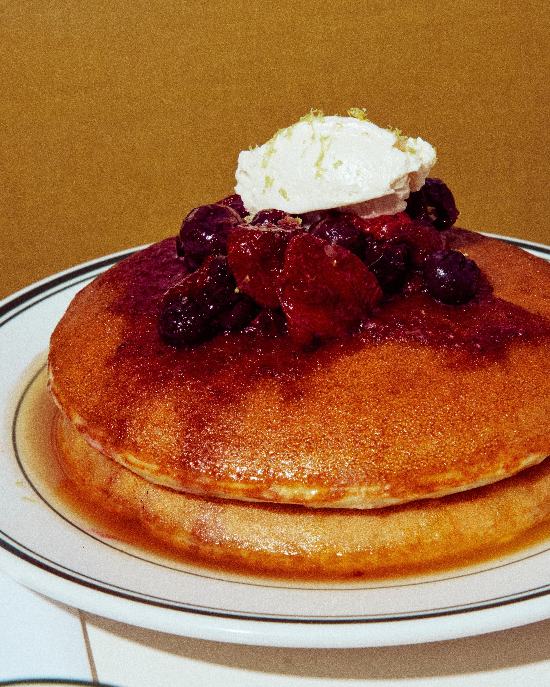 A tall stack of Golden Diner's honey butter pancakes topped with berry compote and whipped butter, syrup pooling on a green-rimmed plate