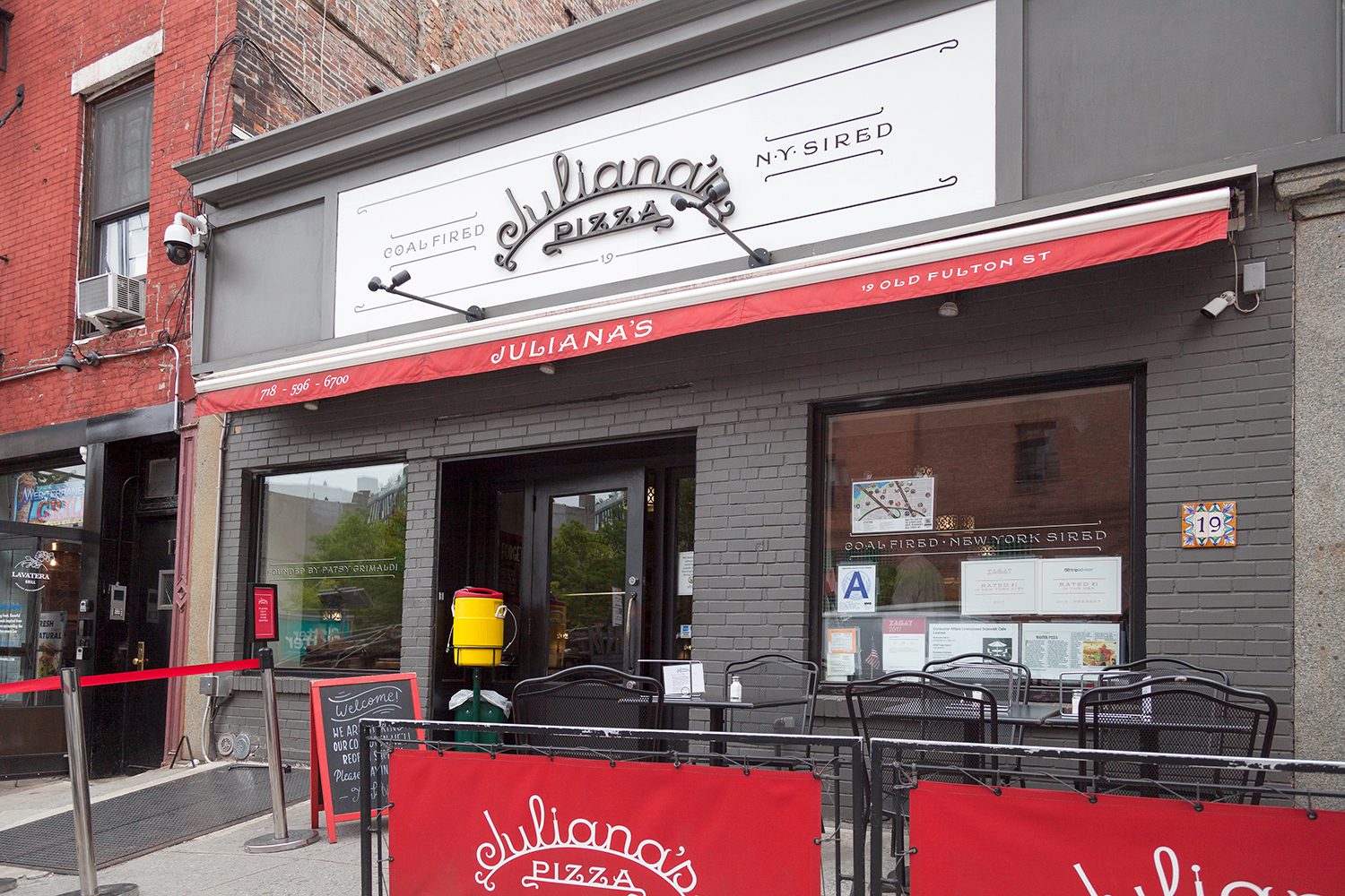 The grey storefront of Juliana's Pizza at 19 Old Fulton Street, with its white script sign, red awning trim and red Juliana's banners around sidewalk tables under the Brooklyn Bridge.